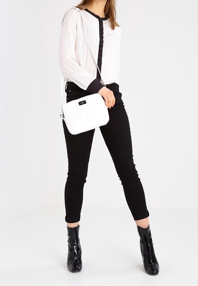 Kvinna i svarta anklarboots, svarta byxor, vit blus med svarta detaljer, som bär en vit axelväska mot en vit bakgrund.