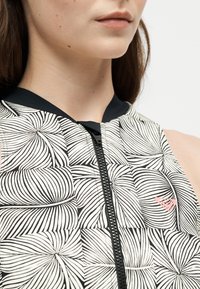 Gros plan d'une femme portant un haut à motifs noir et blanc avec une fermeture éclair frontale et un petit logo cœur rose sur la poitrine.