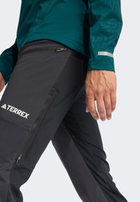 Pantalons TERREX noirs avec poche latérale zippée, associés à un haut à manches longues teal comportant un motif texturé et des accents réfléchissants.