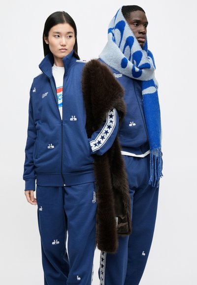 Två personer i matchande blåa träningsdräkter med vita stjärnstripes och kaninlogotyper, en med en stor blå och grå halsduk och brun pälsjacka.