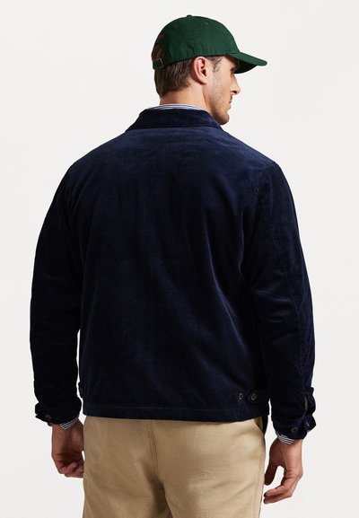 Laivynavių spalvos kordurojinis švarkas su apykakle, mygtukų rankovėmis ir šoninėmis mygtukų detalėmis. Derinamas su smėlio spalvos kelnėmis. Audinys rodo matomą perbrauktą struktūrą.