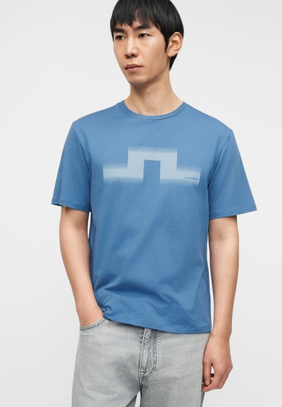 Ανοιχτό μπλε βαμβακερό μπλουζάκι με διακριτικό γραφικό σχέδιο στο στήθος. Κοντά μανίκια, στρογγυλή λαιμόκοψη και άνετη εφαρμογή.