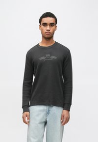 Fiatal férfi fekete, texturált hossz�ú ujjú pólóban, melyen finom láng és pajzs motívum látható, világoskék farmernadrággal, fehér háttér előtt állva.