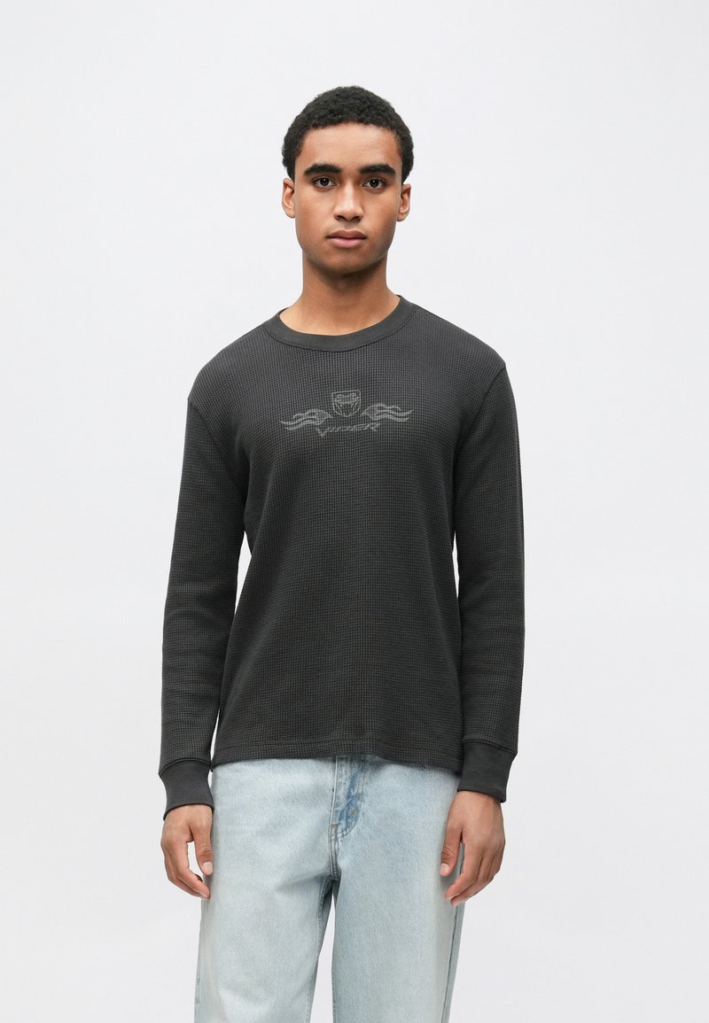 Fiatal férfi fekete, texturált hosszú ujjú pólóban, melyen finom láng és pajzs motívum látható, világoskék farmernadrággal, fehér háttér előtt állva.