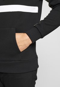 Sudadera negra de mezcla de algodón, con una franja horizontal blanca, puños acanalados y un bolsillo frontal. Textura suave con un diseño ajustado.