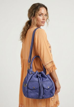 Vrouw met golvend haar in perzikkleurige jurk draagt een grote blauwe leren zak met trekkoord, voorvakken en een lange riem over de schouder.