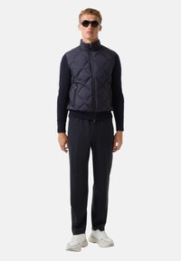 Gilet matelassé bleu marine avec col haut, deux poches zippées sur les côtés, porté par-dessus une chemise à manches longues bleu marine et un pantalon noir avec des baskets.