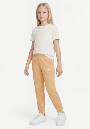 Blondína stojí oblečená v béžovom tričku, hnedých teplákoch Nike a bielych teniskách na bielom pozadí.