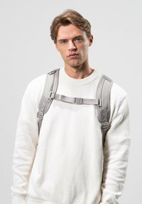 Ryggsäck i ljusgrått tyg med justerbara remmar, med en central spänne och vadderade axelpartier, bärs ovanpå en vit tröja.