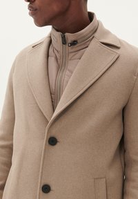 Bruine wollen overjas met een gestructureerde afwerking, voorzien van een notch-kraag, drie knopen en een beige ritsvoering aan de binnenkant.