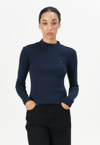 Maglia a maniche lunghe blu navy con un collo alto, design aderente e delicato logo accentato sul petto. Abbinata a pantaloni neri.
