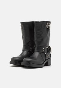 Botas de cuero negras con un diseño hasta la mitad de la pantorrilla, acentuadas con una hebilla circular y un tacón grueso. Textura suave con detalles de correa ajustable.