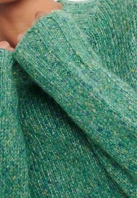 Primer plano de una persona que lleva un suéter de punto verde y moteado, subiendo el cuello hacia su cuello.