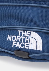 Primo piano di un tessuto blu con il logo bianco ricamato di The North Face e dettagli della tracolla nera.