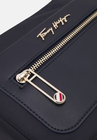 Granatowa torba z złotym zamkiem błyskawicznym i uchwytem, ozdobiona czerwono-biało-niebieskimi paseczkami, z wytłoczonym złotym logo Tommy Hilfiger nad zamkiem.