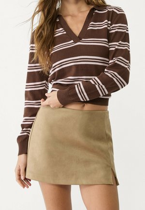 Femme portant un pull à manches longues rayé marron et blanc et une mini-jupe en daim beige avec une petite fente latérale, main reposant près de la taille.