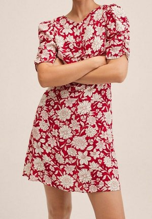 Czerwona kwiatowa sukienka z krótkimi bufkowymi rękawami i marszczeniem, noszona z rękami skrzyżowanymi na piersi, beżowe tło.