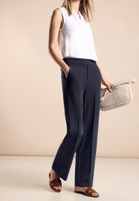 Vit ärmlös topp med V-hals, parat med marinblå vida byxor. Håller en strukturerad beige vävd väska och bär bruna sandaler med remmar.