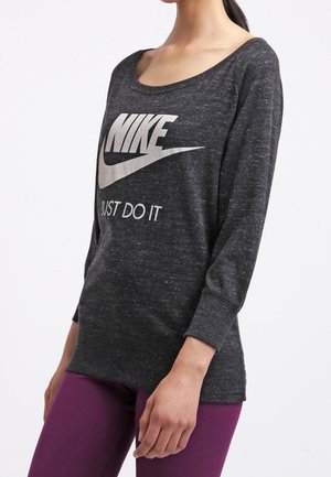 Woman wearing dark gray long-sleeve Nike shirt with "Just Do It" text and purple leggings, standing against a plain white background.
