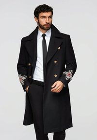 Schwarzer Wollmantel mit doppelt geknöpften goldenen Knöpfen, verziert mit Manschetten mit roten und klaren Edelsteinen. Getragen über einem weißen Hemd und einer schwarzen Krawatte.