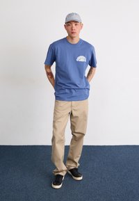 Kék pamut póló fehér kör alakú grafikai logóval, bézs chino nadrág és fekete sneakers fehér talppal. A modellen csíkos sapka van.