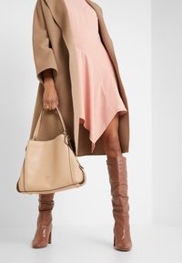 Sac en cuir beige avec une forme souple et structurée, comportant un détail de logo doré et une bordure foncée contrastante. Associé avec une robe rose et un manteau camel.