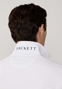 Mann mit dunklem, gewelltem Haar, von hinten gesehen, trägt ein weißes Hemd mit Kragen, auf dem "HACKETT" auf dem Rücken des Kragens gedruckt ist.