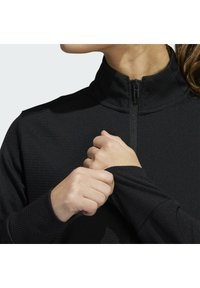 Schwarze Zip-Jacke mit hohem Kragen, strukturiertem Oberflächen und engen Ärmeln. Hände justieren den Bund, was den eng anliegenden, dehnbaren Stoff zur Schau stellt.