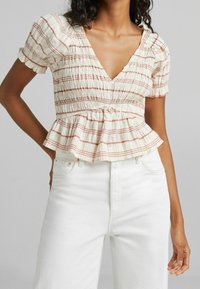 Top bianco con righe orizzontali rosse, corpino arricciato, maniche corte a palloncino e orlo a peplum, abbinato a jeans bianchi a vita alta.