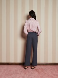 Blusa a maniche lunghe di un rosa chiaro con motivo e colletto, abbinata a pantaloni larghi grigi scuri e tacchi con stampa leopardata, su uno sfondo con parete a righe.