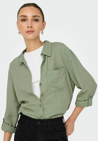 Lichtgroen, knoopshirt met een borstzak, opgerolde mouwen en een zachte textuur, gecombineerd met een witte T-shirt en zwarte spijkerbroek.