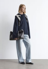 Marineblauer Strickpullover in lockerer Passform, kombiniert mit hellblauen weit geschnittenen Jeans und schwarzen spitzen Ballerinas. Schwarze Lederhandtasche mit gefaltetem Oberteil.