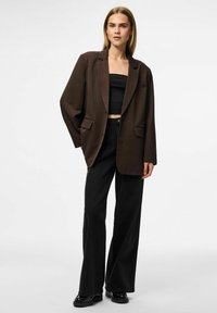 Rjava oversize blazer z enim gumbom, dvema sprednjima žepoma, v kombinaciji s črnimi širokimi hlačami in sijočimi čevlji.