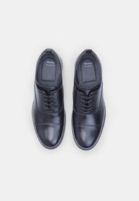 Chaussures habillées en cuir noir avec une finition lisse, design à lacets, bout rond et détails de couture minimaux. Logo de la marque visible à l'intérieur.