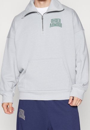 Jasnoszara bluza z suwakiem do ćwiartki z logo "Under Armour" na piersi, w połączeniu z granatowymi spodniami z tym samym logo na lewej udzie.