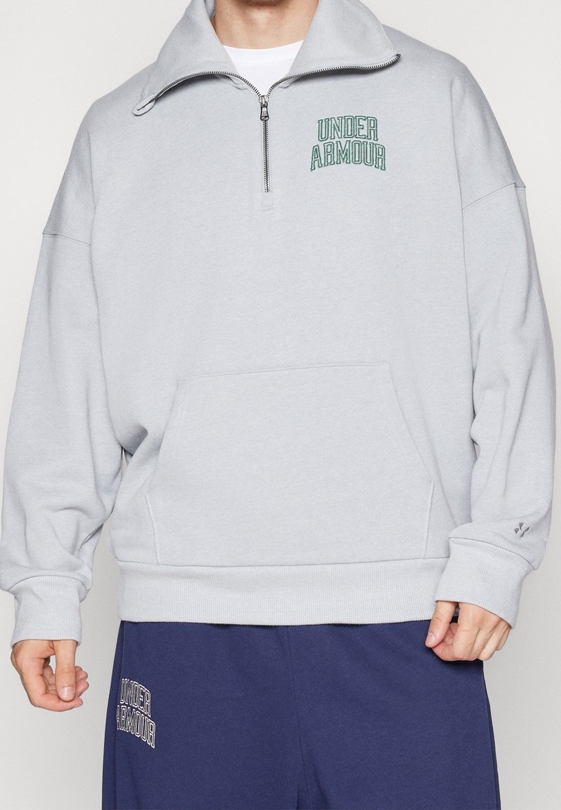 Sweat-shirt gris clair à fermeture éclair quart de zip avec le logo "Under Armour" sur la poitrine, associé à un pantalon bleu marine avec le même logo sur la cuisse gauche.