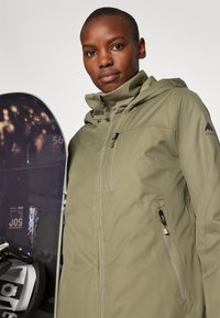 Olivovo zelená nepremokavá bunda s vysokým golierom, zipsovými vreckami a logom na rukáve. V popredí je snowboard s tmavými grafikami.