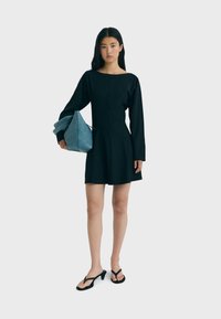 Schwarzes langärmliges Kleid mit Bootsausschnitt, tailliertem Oberteil und ausgestelltem Rock. Das Model hält eine blaue Tasche und trägt schwarze Flip-Flops.