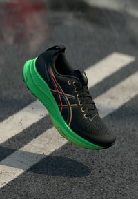 Schwarzer Laufschuh mit strukturiertem Obermaterial, grüner Sohle und roten Akzenten. Verfügt über Schnürsenkel und Branding auf der Zunge, das ein leichtes Design hervorhebt.