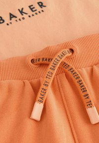 Tejido de sudadera en color melocotón con puños acanalados. Presenta un cordón con marca, acabados de costura plana y una textura suave.