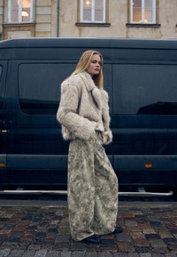 Beżowa krótka kurtka z sztucznego futra, szerokie oliwkowe spodnie w wzory, czarne buty, na tle ciemnego vana i kamiennej nawierzchni.