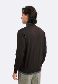 Cardigan marrone scuro con collo e polsini a coste, maglia strutturata, design aderente e colore uniforme. Indossato sopra una camicia blu con motivo.