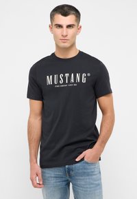 Melna kokvilnas T-krekls ar balto "MUSTANG" logo, ar klasisku apaļu apkakli un īsām piedurknēm, apvienots ar ziliem džinsu biksēm.