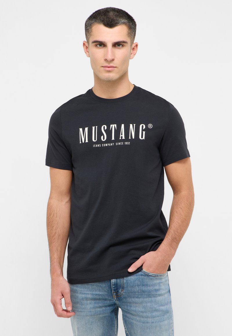 Melna kokvilnas T-krekls ar balto "MUSTANG" logo, ar klasisku apaļu apkakli un īsām piedurknēm, apvienots ar ziliem džinsu biksēm.