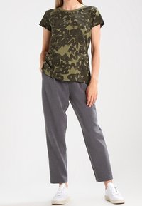 Olivově zelené tričko s maskáčovým vzorem, v kombinaci se šedými volnými kalhotami. Modelka má na sobě bílé tenisky a pózuje s jednou rukou na bocích.