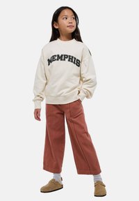 Une jeune fille portant un sweat-shirt crème "Memphis", un pantalon large couleur rouille, des chaussettes grises et des chaussures slip-on beige, se tenant avec une main dans sa poche.