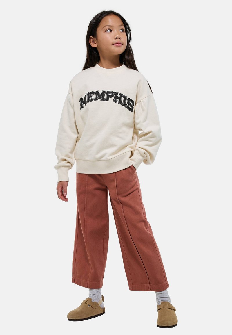 Une jeune fille portant un sweat-shirt crème "Memphis", un pantalon large couleur rouille, des chaussettes grises et des chaussures slip-on beige, se tenant avec une main dans sa poche.