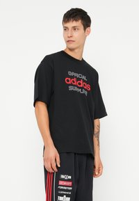 Melnas īsām piedurknēm t-krekls ar sarkaniem un baltajiem uzrakstiem "OFFICIAL adidas SUPPLIER". Vienkāršs dizains ar apļveida kakla izgriezumu un brīvu piegriezumu.