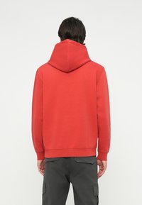 Červená mikina s kapucňou vyrobená z mäkkej tkaniny, s veľkou kapucňou, rebrovanými manžetami a jednofarebným dizajnom. Pohľad z chrbta, žiadne viditeľné logá ani vzory.