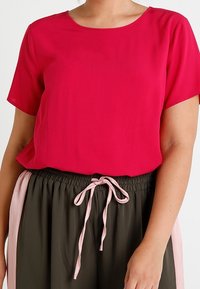 Blusa rossa a maniche corte con scollatura tonda, abbinata a pantaloni verde oliva con vita elasticizzata, strisce laterali rosa e stringhe.